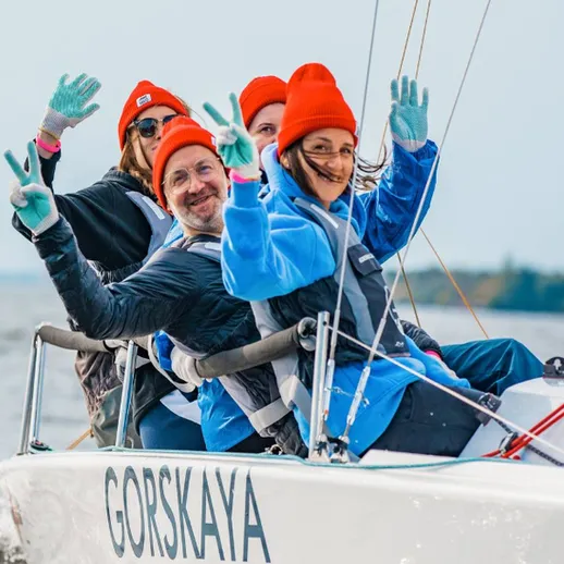 Команда на лодке с парусами радуется солнечному дню на водоеме, одетая в яркие шапки, береговая линия на фоне