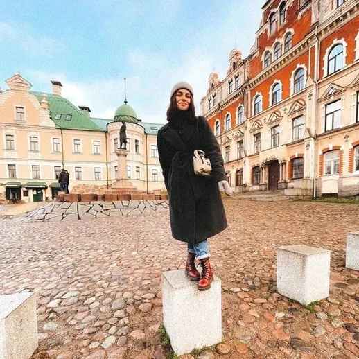 Девушка в черном пальто стоит на постаменте на площади с историческими зданиями в холодный день