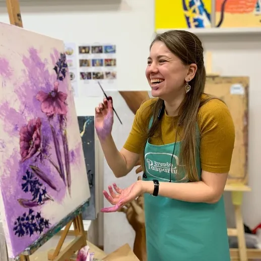 Молодая художница создает картину с фиолетовыми цветами, улыбаясь в художественной мастерской в дневное время