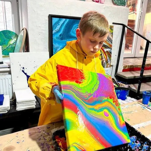 Мальчик создает яркую абстрактную картину в мастерской, окруженный красками и материалами, время – дневные часы