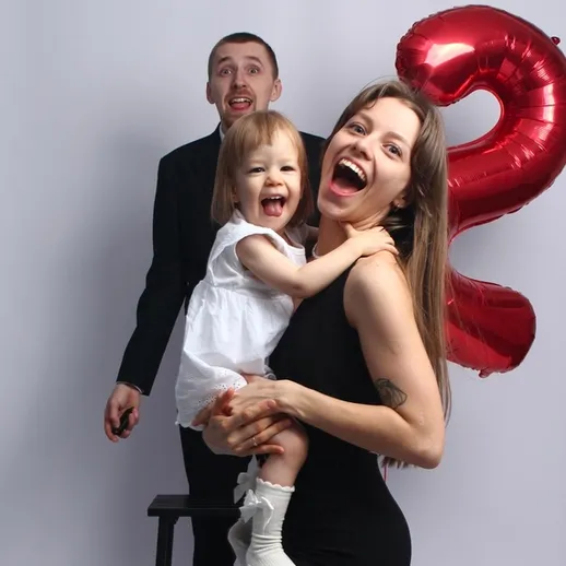 Счастливая семья отмечает день рождения ребёнка с шариком в форме цифры два на фоне нейтральной стены