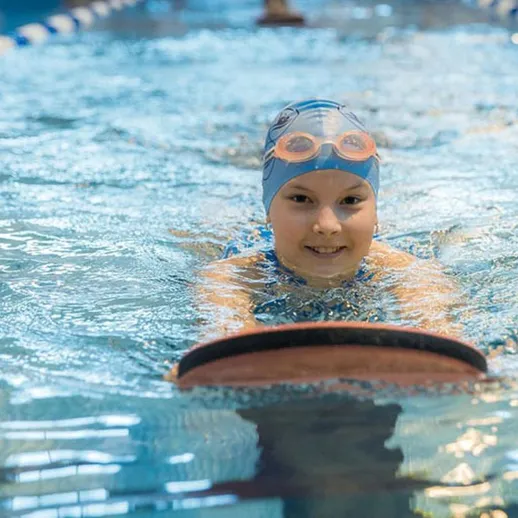 Мальчик в шапочке плавает в бассейне, используя доску для плавания в спортивном зале в дневное время