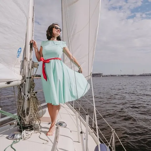 Женщина в светлом платье наслаждается ветром на парусной лодке у берега в солнечный день