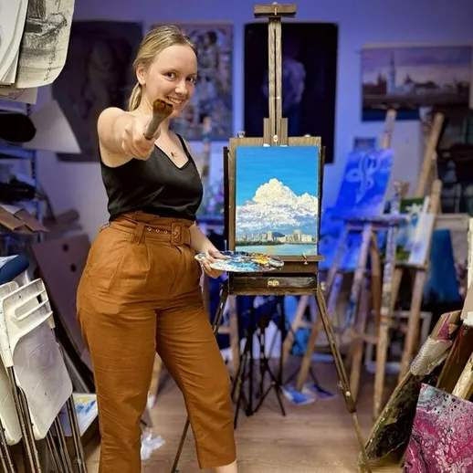 Молодая художница позирует с кистью перед своим пейзажным холстом в художественной студии