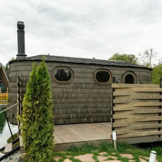 Уютный домик с круглым окном у водоема в загородном комплексе, окруженный зеленью и расположенный вдали от городской суеты