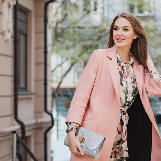 Молодая женщина на улице в стильном розовом пальто и цветной блузке позирует на фоне архитектуры города весной