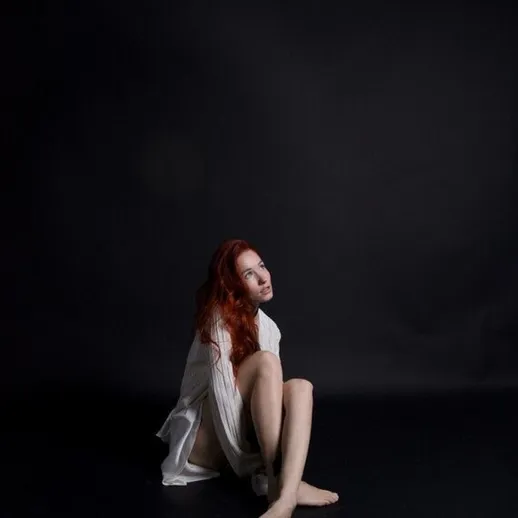 Молодая женщина сидит на полу в пустом темном помещении и смотрит в камеру с загадочной улыбкой