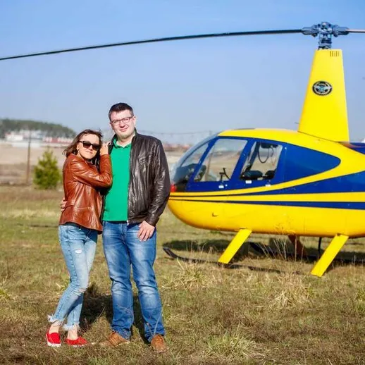 Пара с улыбками позирует рядом с желтым вертолетом на фоне чистого неба и зелёной травы в солнечный день