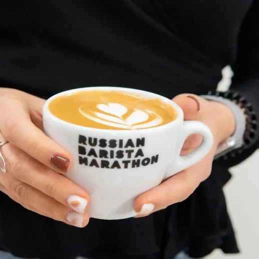 Участница марафона бариста показывает латте-арт в чашке с надписью
