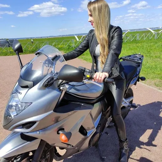 Молодая женщина в кожаном костюме управляет мотоциклом на фоне зелёного поля в ясный день