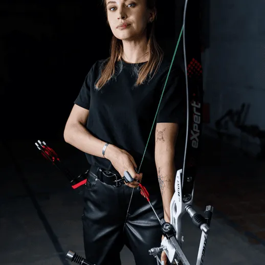 Архерша в черной одежде позирует с луком и стрелами в помещение для стрельбы с луком