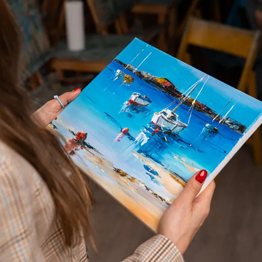 Женщина рассматривает картину с морским пейзажем и яхтами в уютной студии в солнечный день