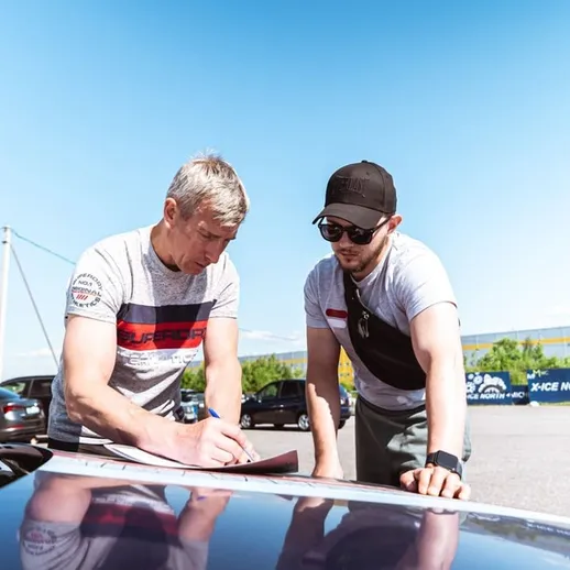 Два человека обсуждают детали на капоте автомобиля под ясным небом в дневное время