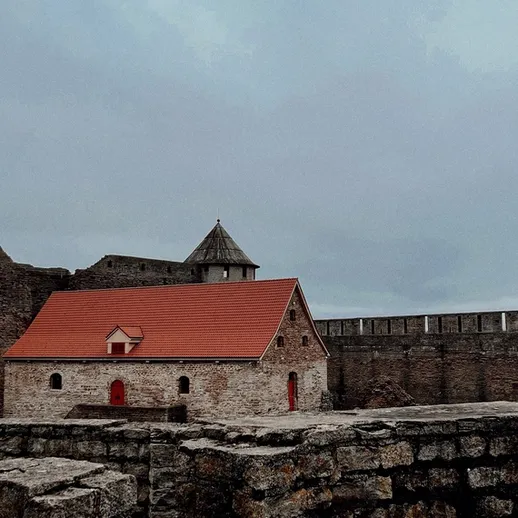 Каменный замок с красной крышей на фоне серого неба и крепостной стены в ясный день