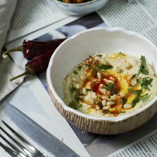 Красивый блюдо хумуса с зеленью, семенами и специями на столе с газетой и прибором