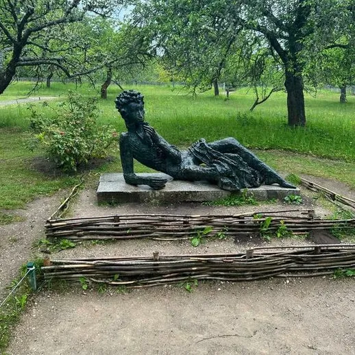 Скульптура женщины, лежащей на постаменте в парке, окруженном зелеными деревьями, в ясный летний день