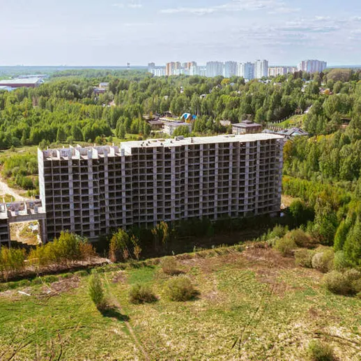 Заброшенное здание в лесу на фоне современного города, окруженное зелеными деревьями под ясным небом