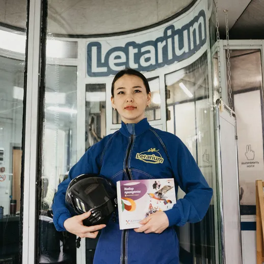 Молодая женщина в синих комбинезоне и шлеме стоит у входа в летарий, держа в руках сертификат о прохождении курса