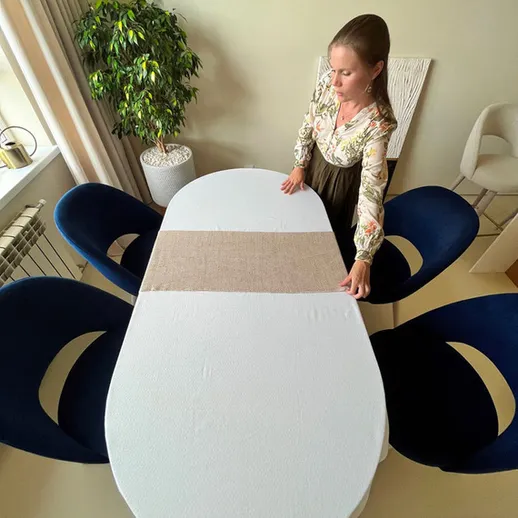 Женщина накрывает стол белой скатертью в светлой комнате с современными стульями