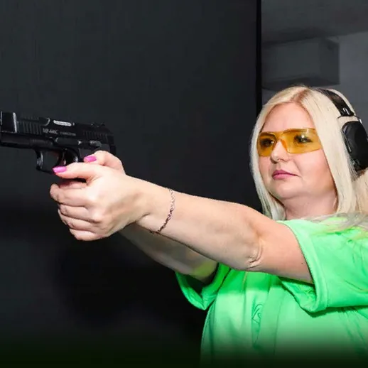 Женщина стреляет из пистолета в тире в зеленой футболке с защитными очками на фоне темной стены