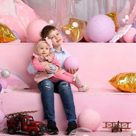 Дети на фоне розовых подушек и декораций, малышка в платье, мальчик с игрушками на ступенях в праздничной обстановке