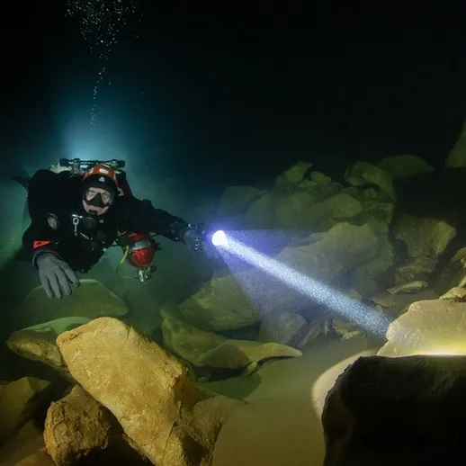 Дайвер исследует подводные камни в темной пещере с мощным фонарем в спокойных водах