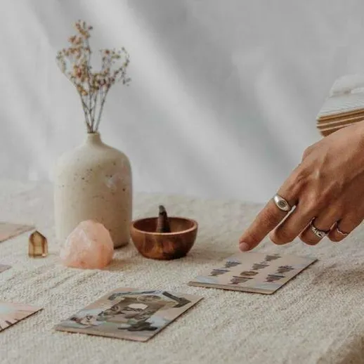 Руки человека указывают на карты оракула на столе в домашней обстановке, окруженные цветами и камнями в мягком свете