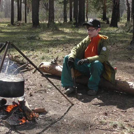 Мальчик готовит еду на костре в лесу, наслаждаясь природой и спокойствием в ясный день