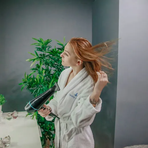 Девушка с феном укладывает волосы в ванной комнате с растениями и зеркалом в вечернее время