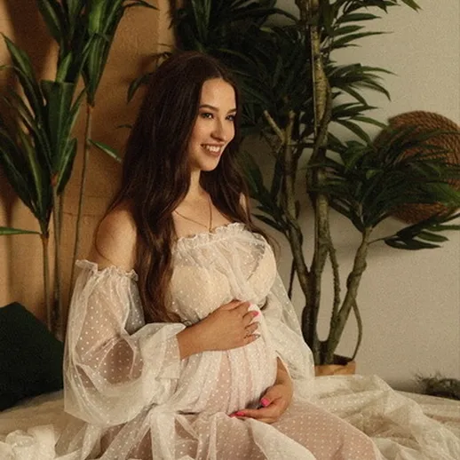 Беременная женщина в белом платье сидит на диване в окружении растений в уютной комнате