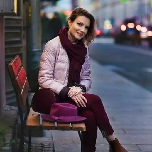 Девушка в розовой куртке и бордовом шарфе, сидит на скамейке в городе в вечернее время