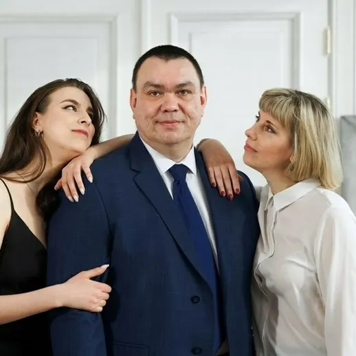 Две женщины обнимают мужчину в деловом костюме в светлом помещении на фоне абстрактного искусства