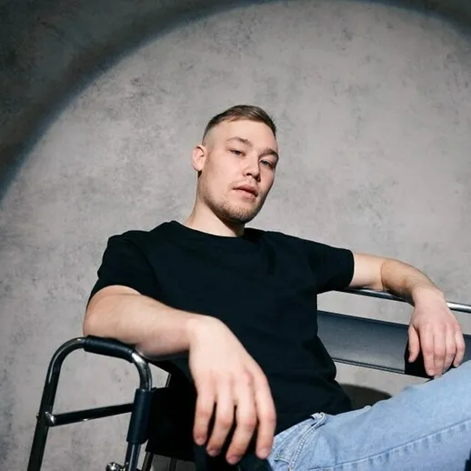 Молодой человек сидит на инвалидной коляске, позируя с уверенной мимикой в нейтральной обстановке