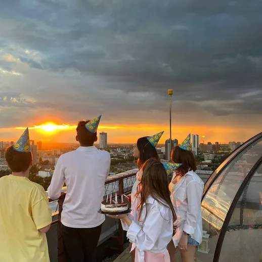 Друзья встречают закат на крыше, празднуя день рождения с тортом и праздничными шапочками в городской атмосфере