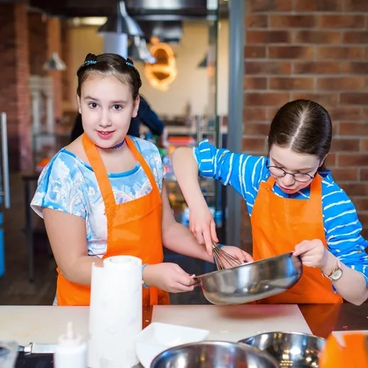 Две девочки в оранжевых фартугах готовят блюдо на кулинарном мастер-классе в уютной студии