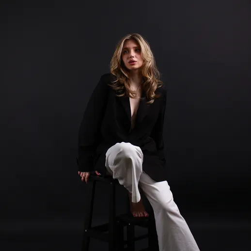 Женщина в черном пиджаке и белых брюках сидит на стуле на черном фоне в студии