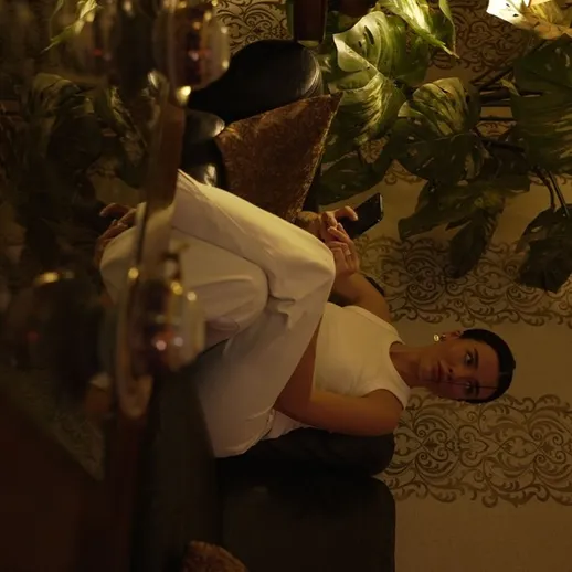 Женщина в белом платье сидит на диване среди зелёных растений в уютной комнате вечерней атмосферой