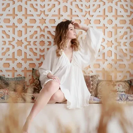 Молодая женщина в белом халате сидит на диване у стены с орнаментом в уютном интерьере