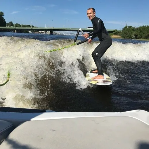Молодой человек серфит на волне, созданной моторной лодкой, на реке в солнечный день
