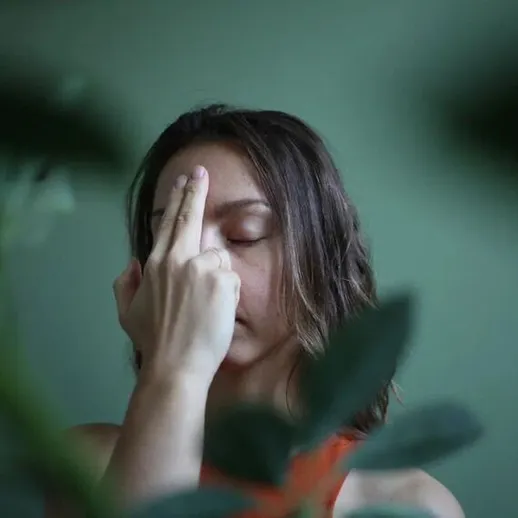 Женщина с закрытыми глазами и рукой на лбу на фоне растений в помещении