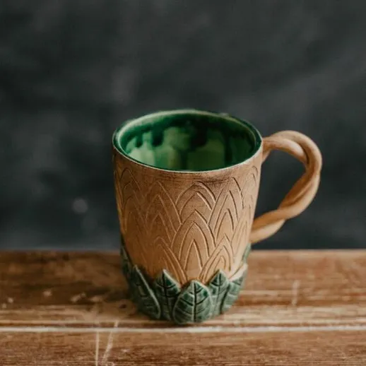 Керамическая кружка с рельефным узором и зелеными деталями на деревянной поверхности в теплых тонах