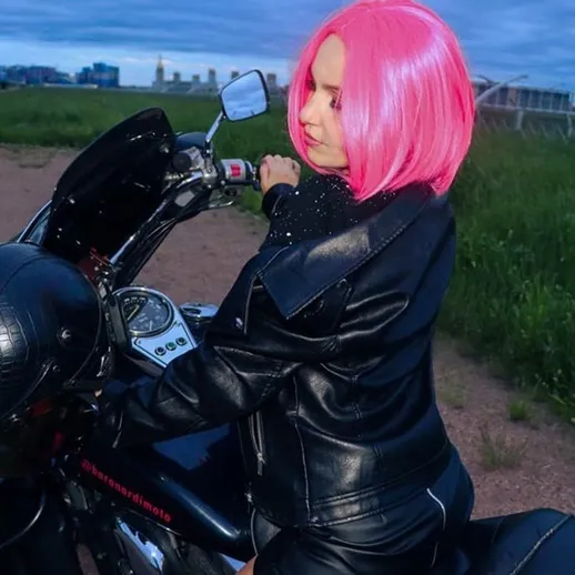 Ярко-розововолосая женщина в кожаном костюме на мотоцикле на фоне вечернего неба