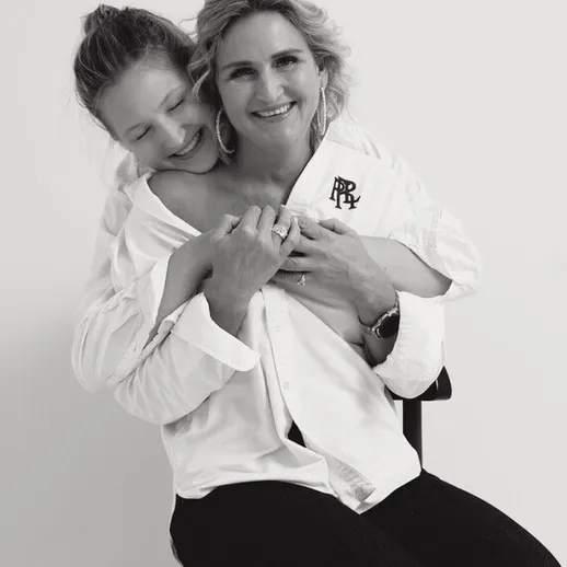 Две женщины обнимаются, позируя в белых рубашках на фоне нейтральной стены