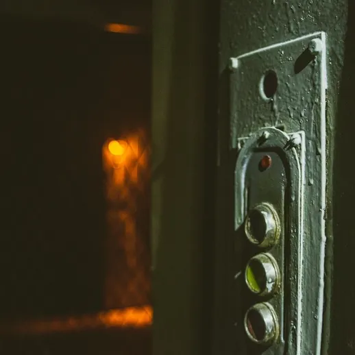 Старая дверь с кнопками вызова лифта, освещённая ненавязчивым светом в тёмном коридоре здания