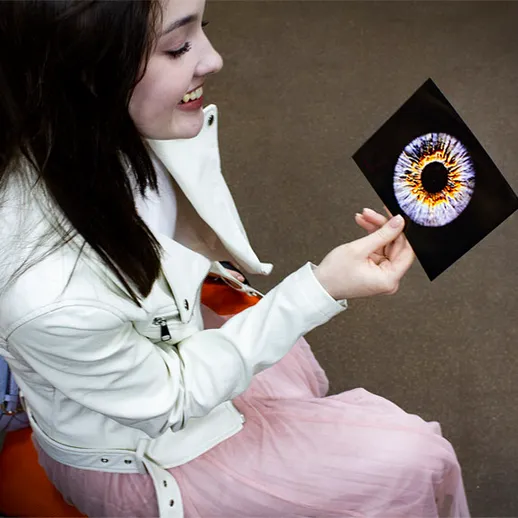 Девушка в белом пиджаке держит изображение глаза на черном фоне в помещении с современным интерьером