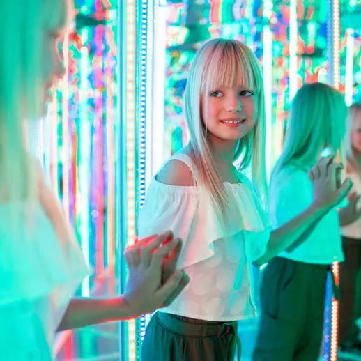 Девочка с длинными светлыми волосами смотрит на свое отражение в зеркалах, окруженная красочными огнями в развлекательном центре