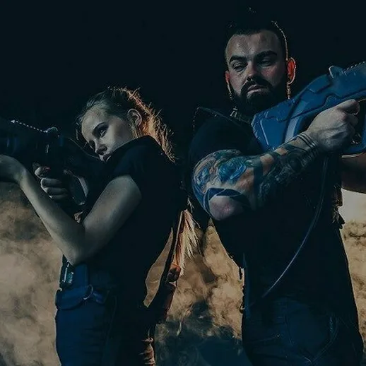 Два человека в черной одежде держат игрушечные пушки на фоне дыма, будто готовясь к игре или тренировке