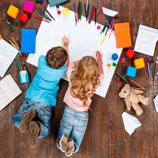 Два ребенка рисуют на большом листе бумаги, окруженные красками и канцелярскими принадлежностями на деревянном полу