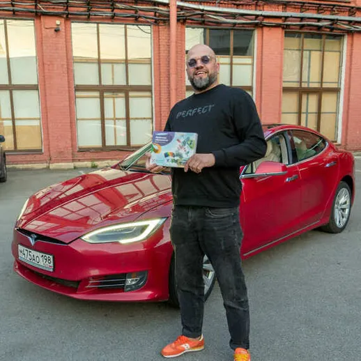 Мужчина стоит рядом с красной машиной в городе, держа в руках книгу, на улице в солнечный день