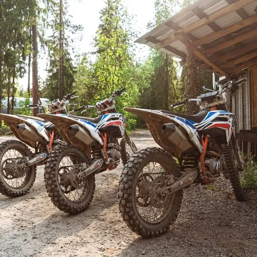Три мотокросса стоят рядом с деревянным зданием на фоне зелёных деревьев в солнечный день